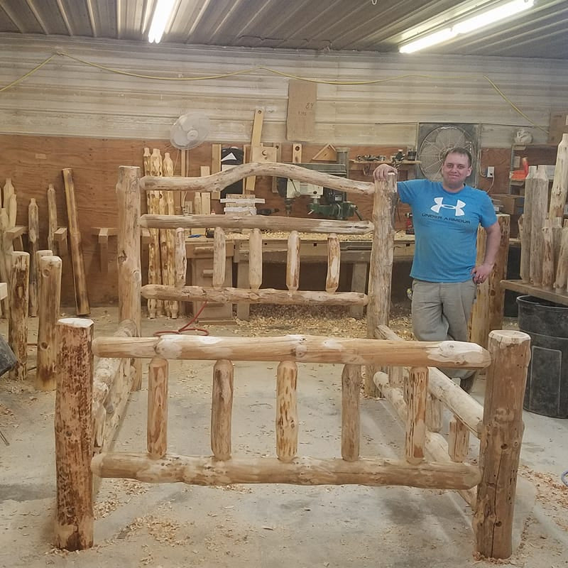 Man standing next to a wooden bed frame in a workshop.